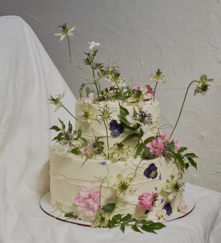 Two-tiered white cake decorated with fresh wildflowers and greenery against white backdrop.