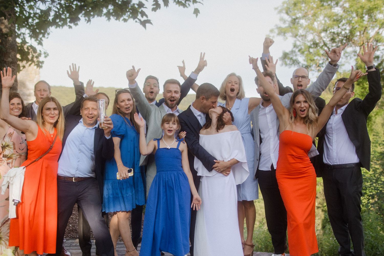 Wedding party celebrates with raised hands and bright attire against a natural mountain backdrop.