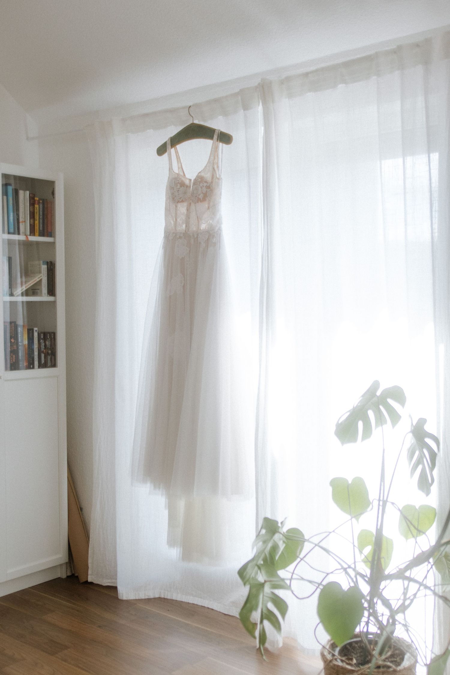 Delicate white wedding dress hanging by window with houseplants and bookshelf in bright room.