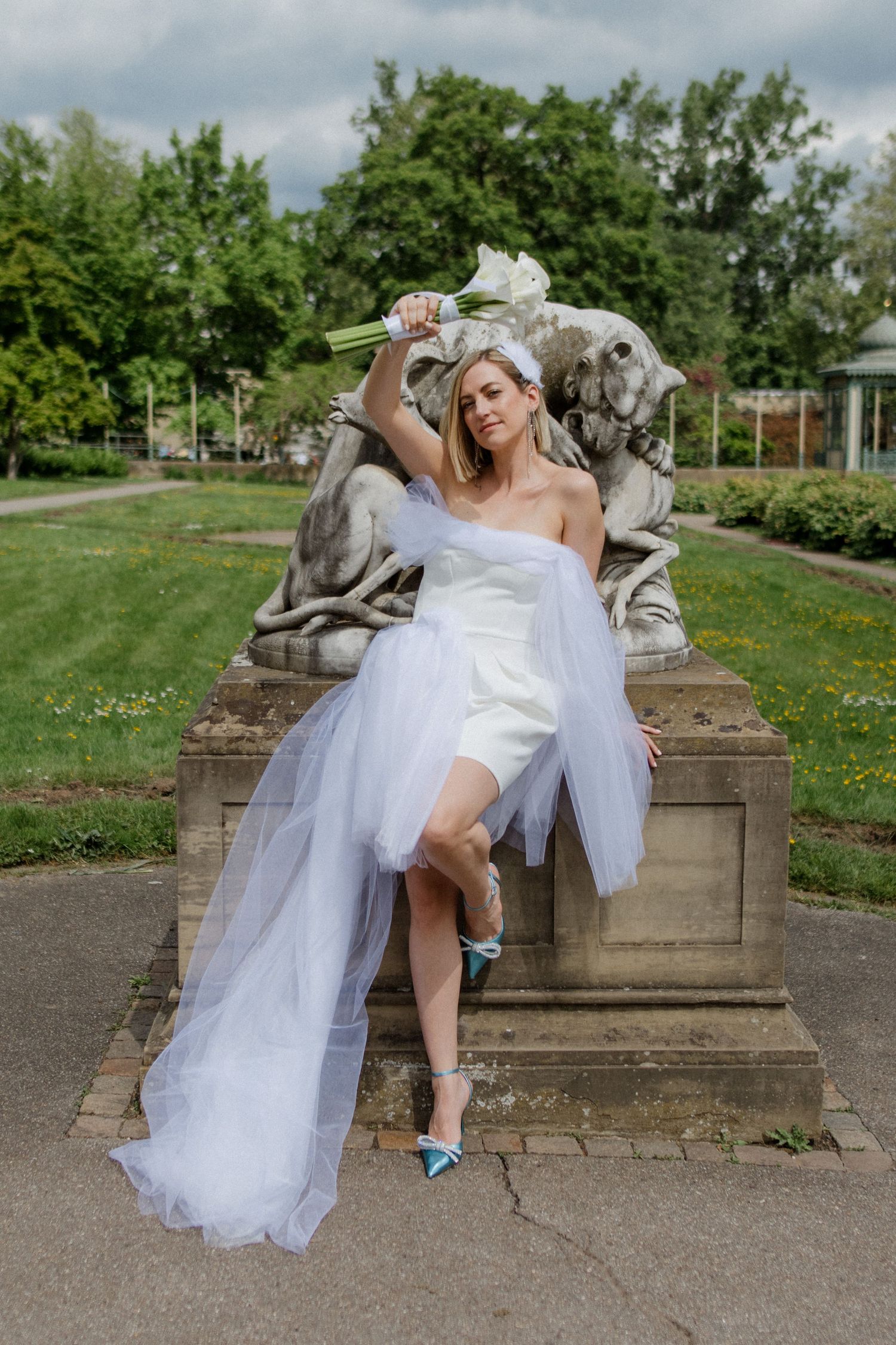 Bride in modern short dress poses elegantly on stone pedestal with calla lily bouquet.
