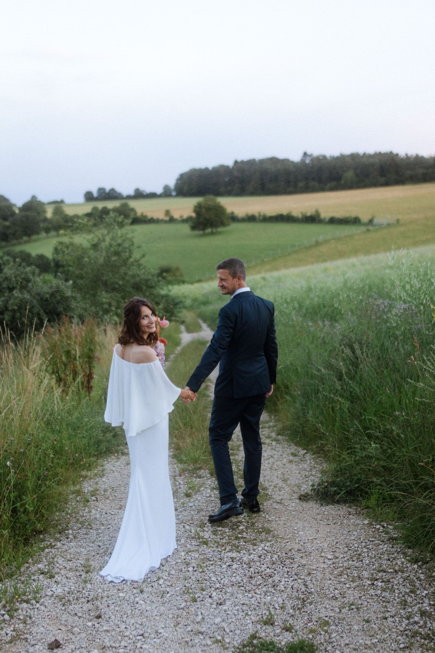 A couple in wedding attire walking hand in hand down a scenic gravel path.