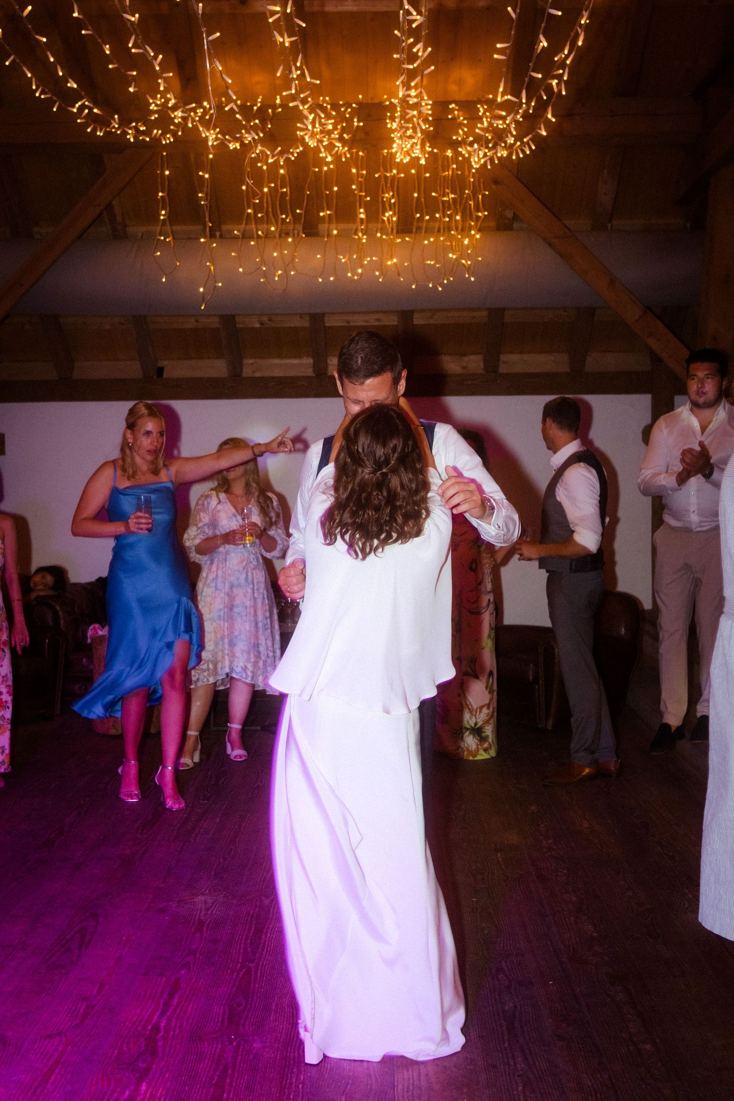 Wedding guests dancing under decorative string lights with purple lighting effects.