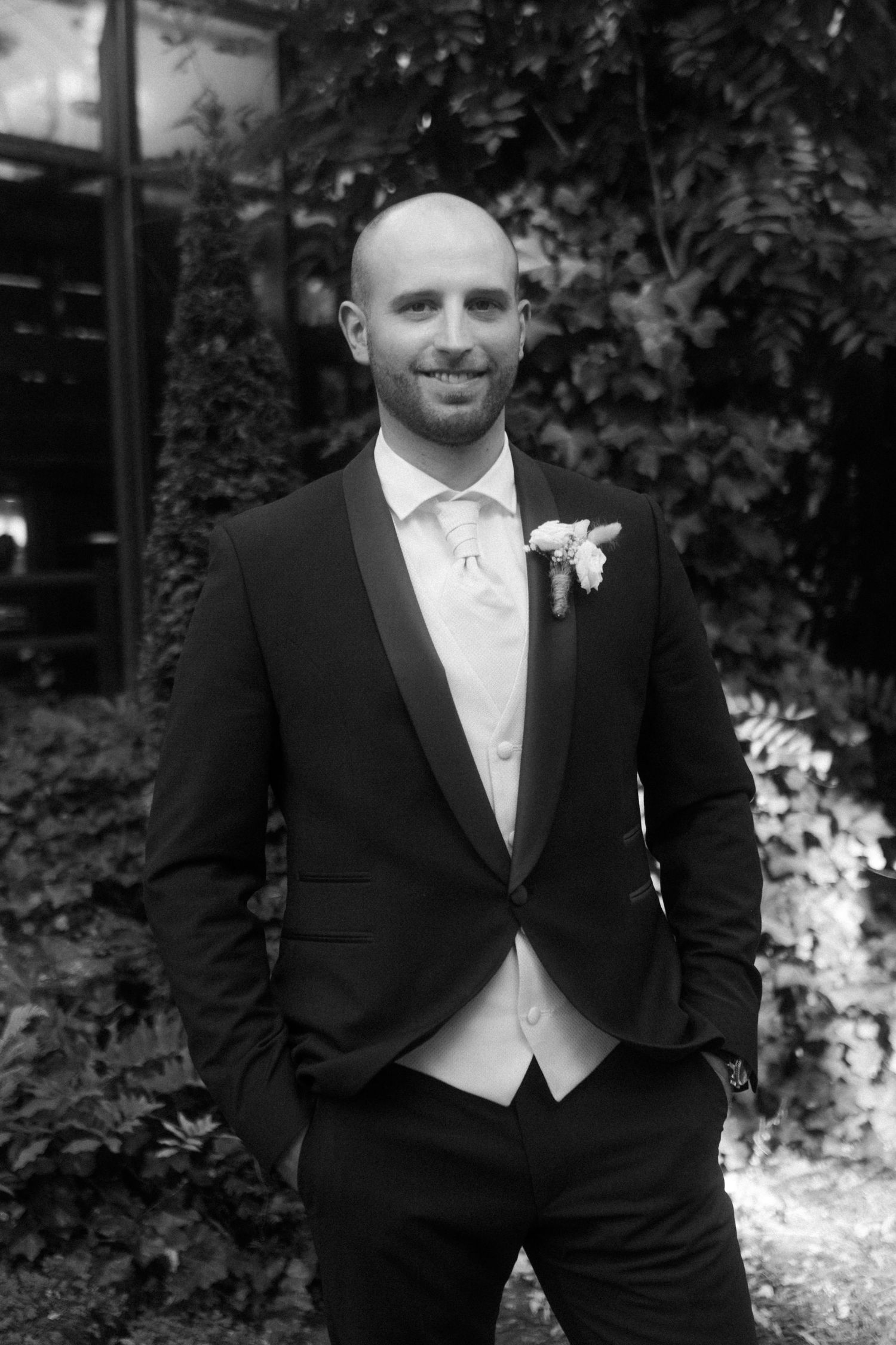 A black and white portrait of a groom in formal wedding attire smiling at the camera.