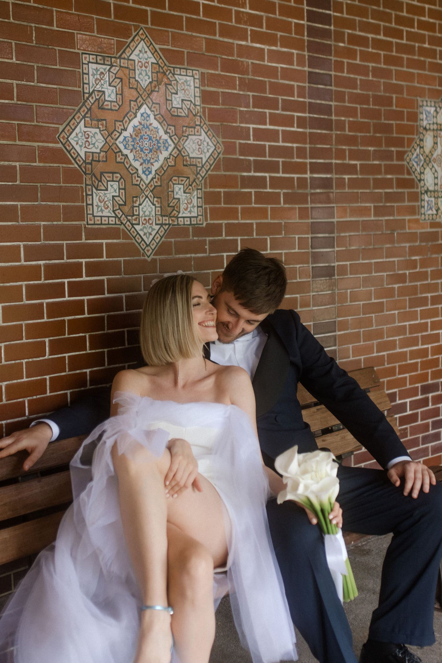 Wedding couple sharing an intimate moment on wooden bench against decorative brick wall.