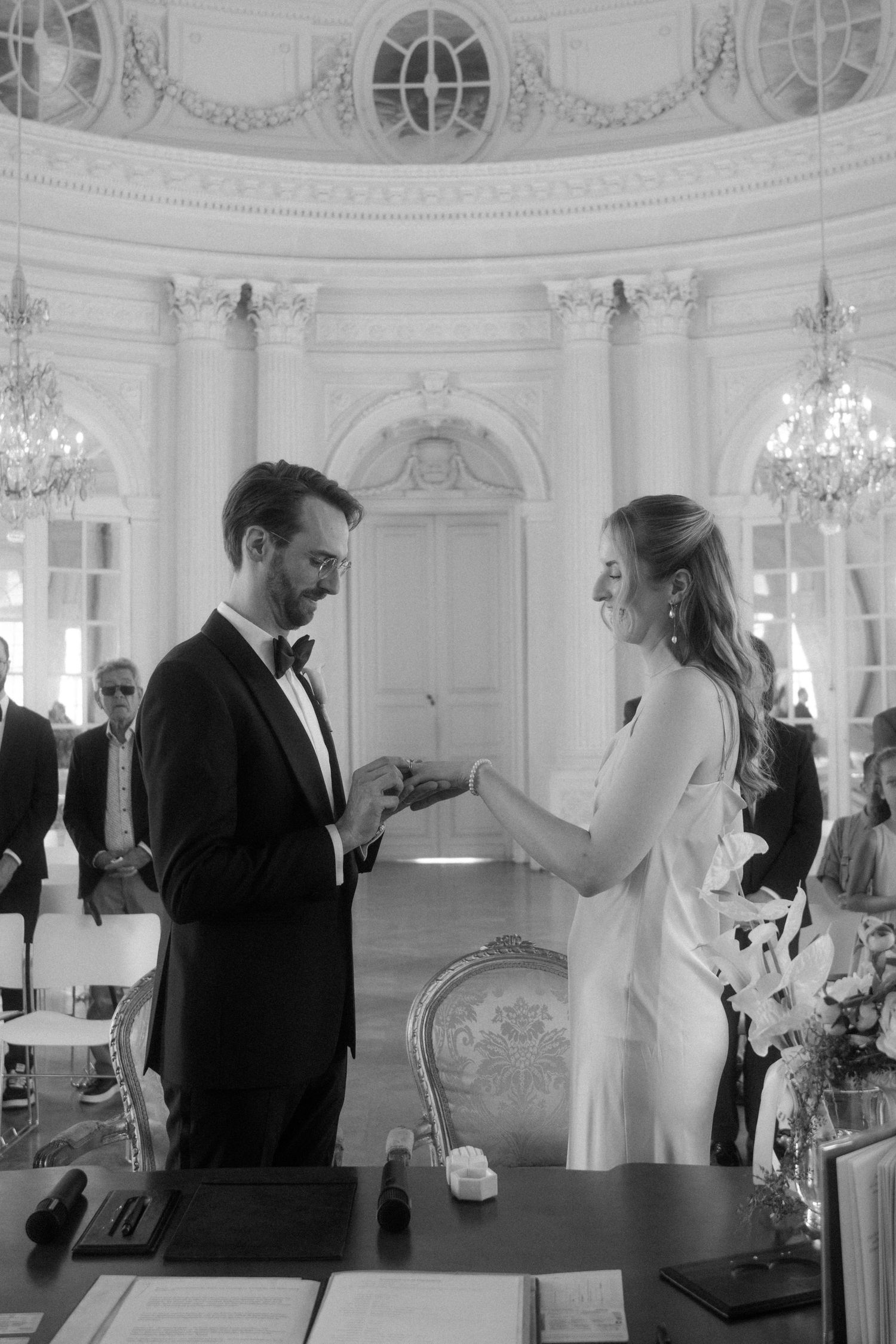 A couple shares an intimate moment in an elegant white ballroom with ornate architectural details and chandeliers.