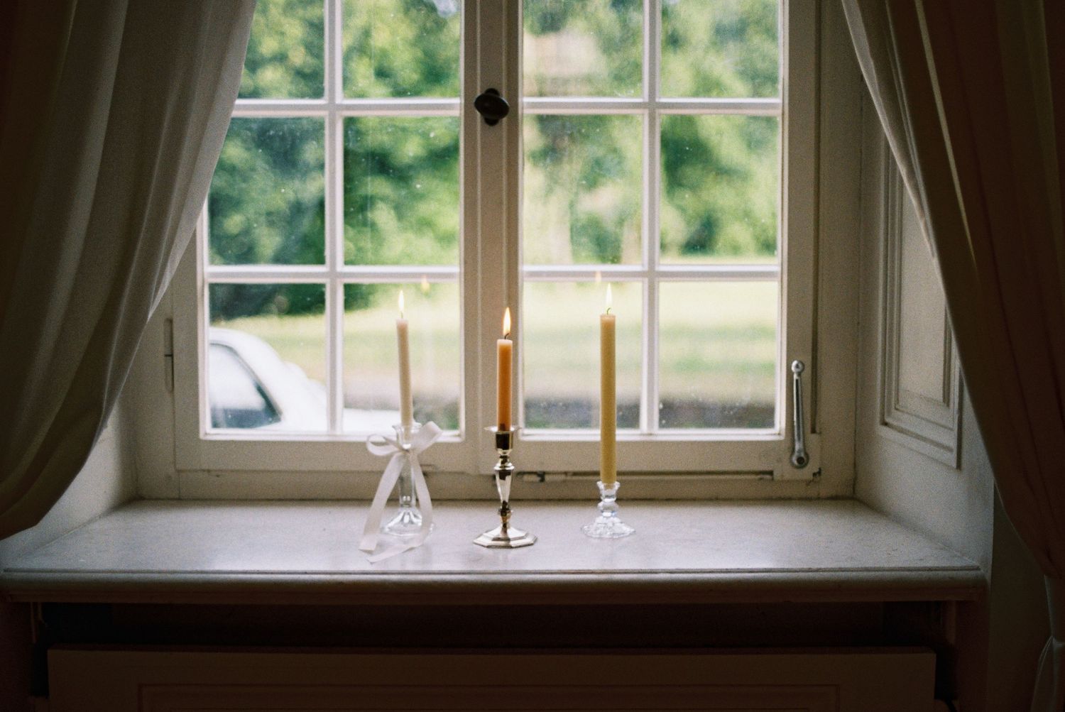 A rustic window sill with candlesticks against a bright window overlooking green countryside.