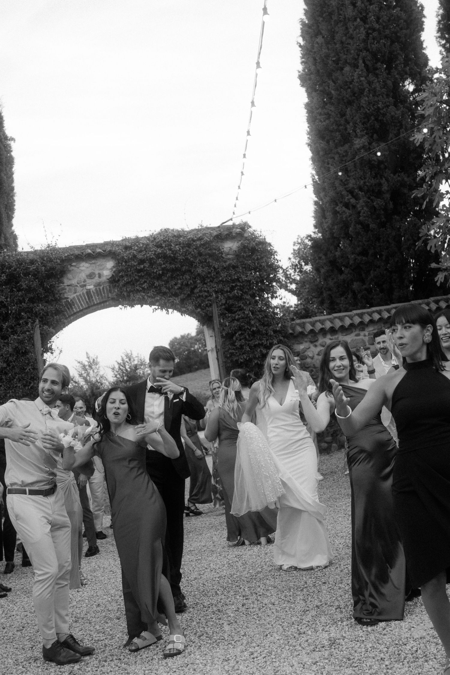 Black and white photo of wedding guests dancing in formation at outdoor reception.
