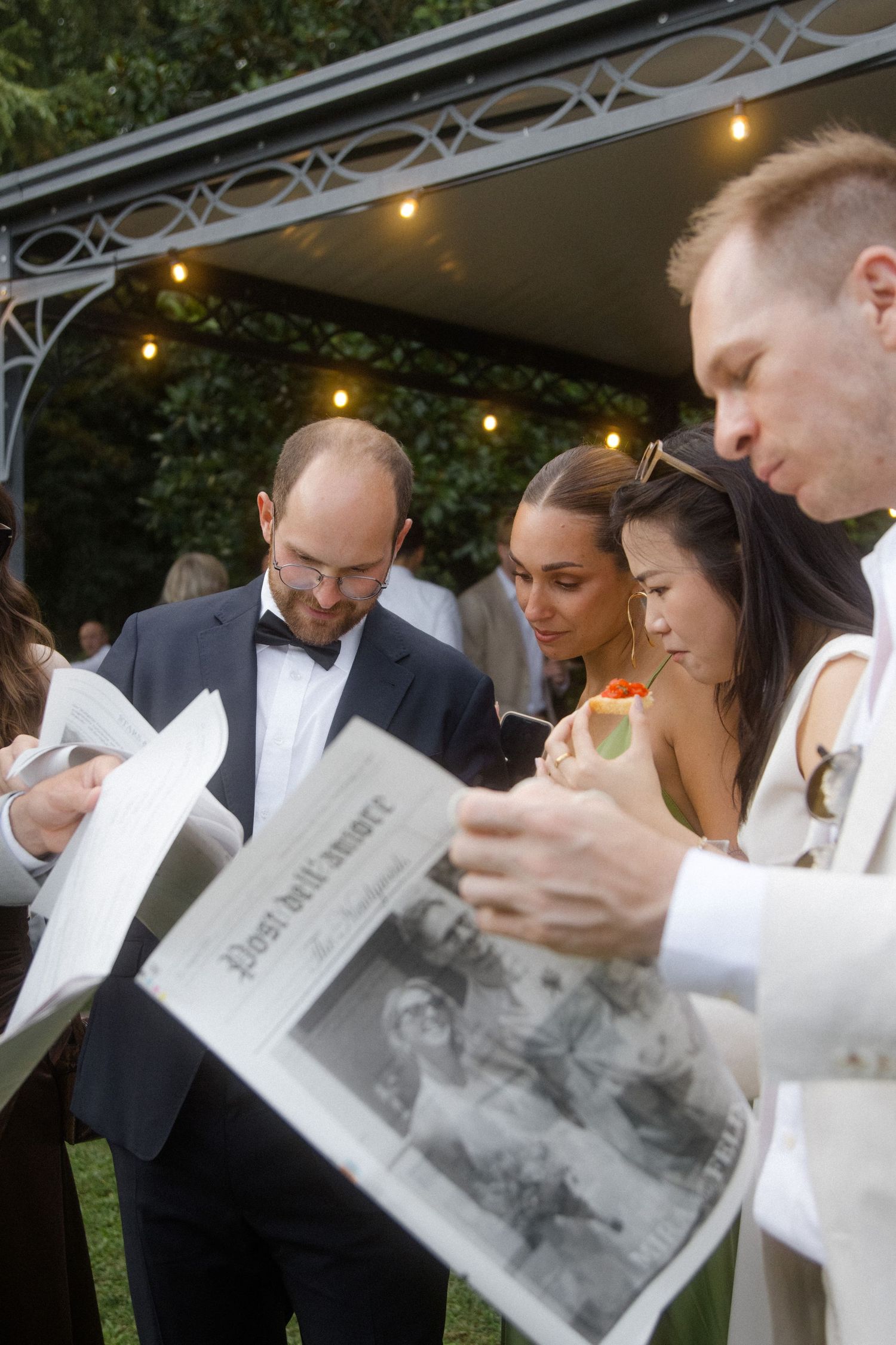 Wedding guests reading programs together under string lights at an outdoor evening ceremony.