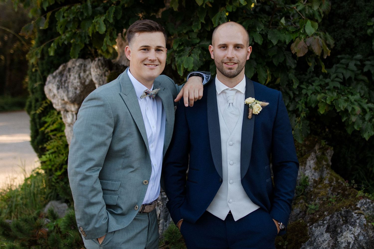 Two men in formal suits posing together against a green garden backdrop.