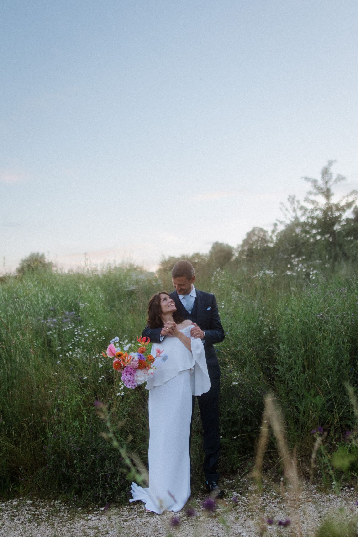 A bride in a sleek white dress holds colorful flowers while being embraced by her groom outdoors.