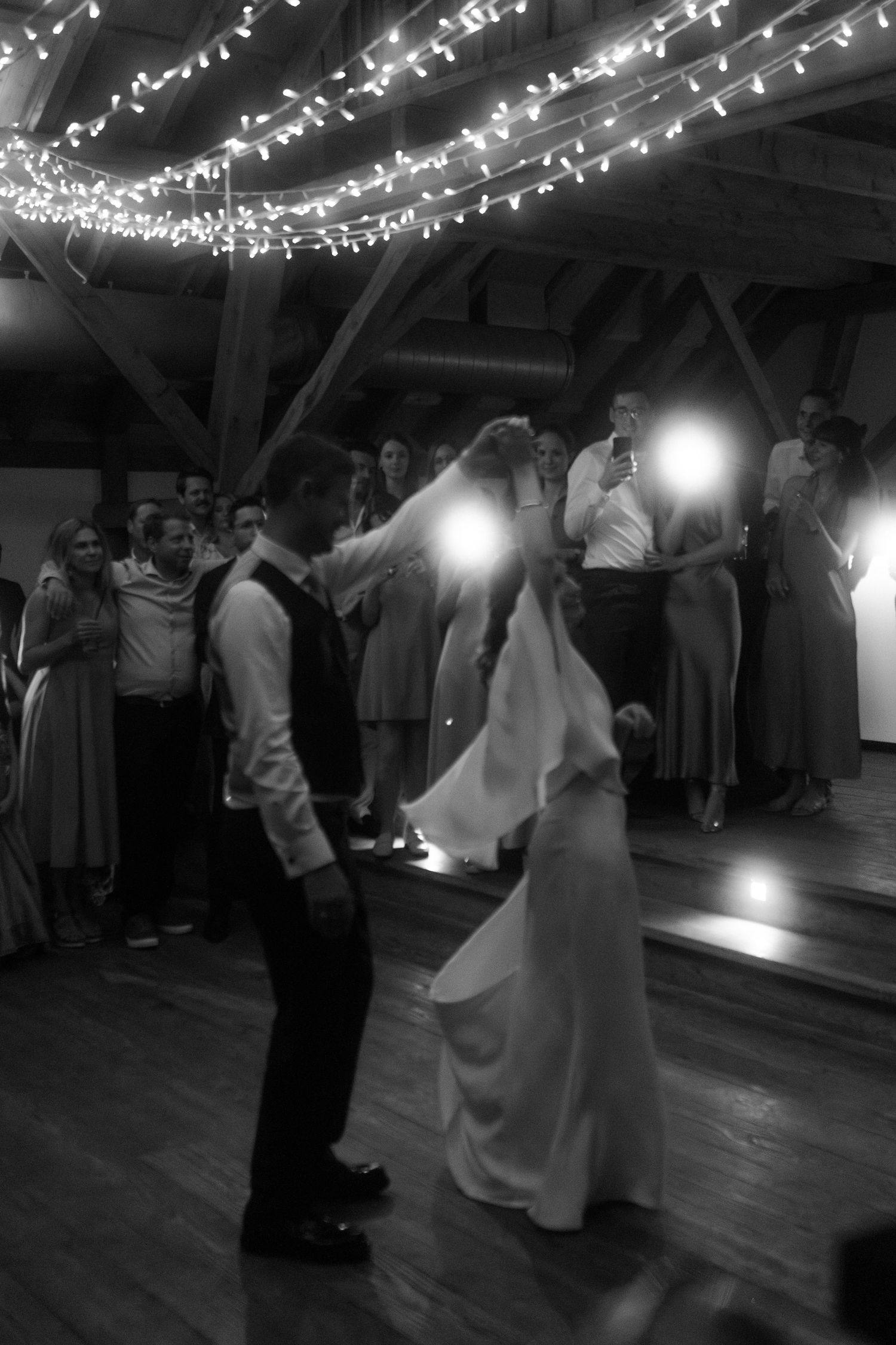 Black and white photo of a dance at a wedding reception with fairy lights overhead.