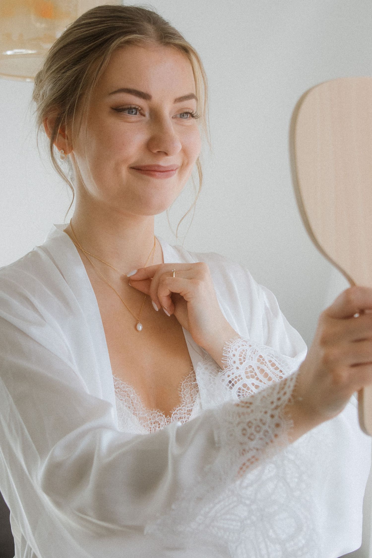 Person in white lace robe looking at hand mirror with soft natural lighting.