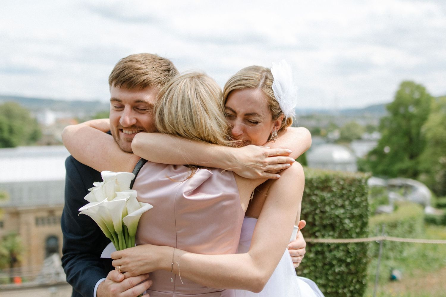 Three people share an emotional group hug during a wedding celebration with scenic hills in the background.