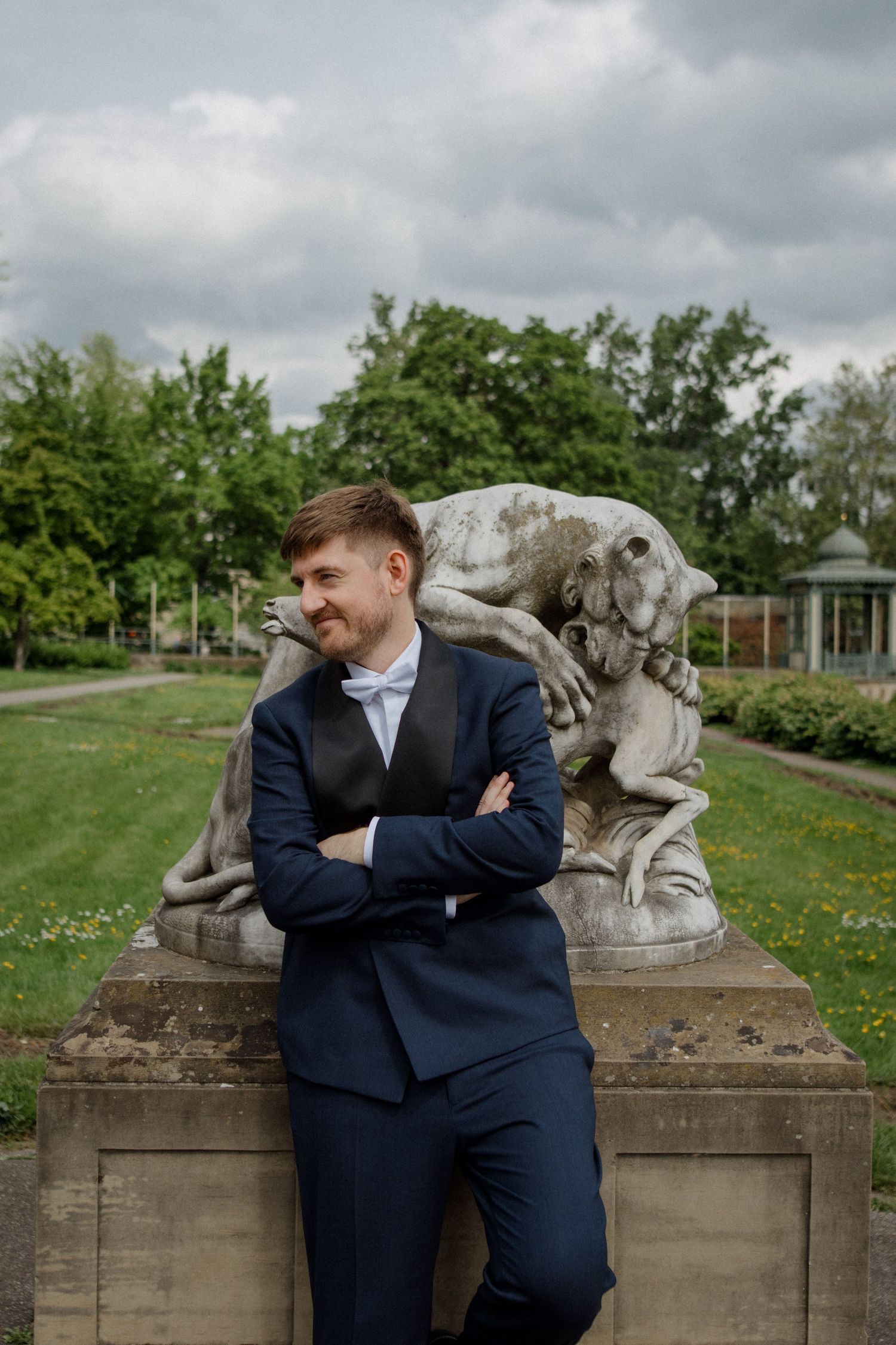 Groom in navy suit poses confidently against stone garden statue under cloudy sky.