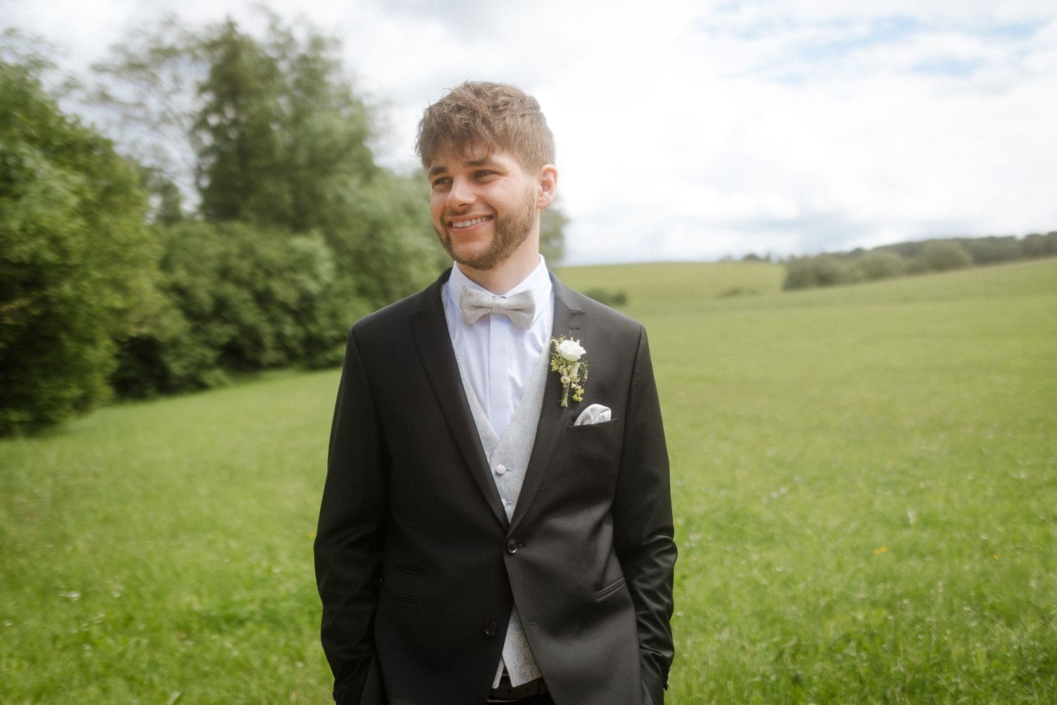 A groom in a black tuxedo and light blue bowtie smiles while standing in a green field on his wedding day.
