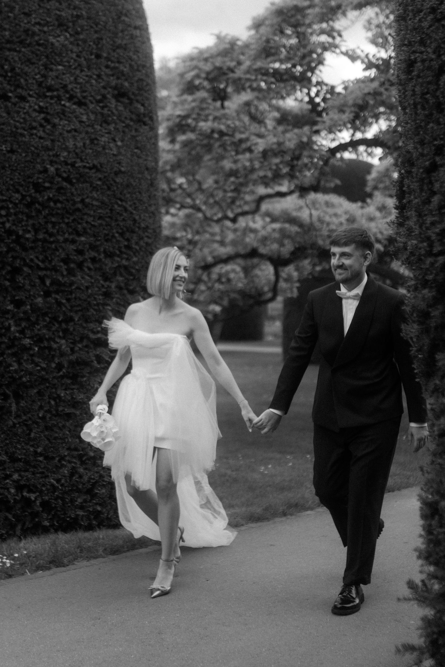 A black and white photo of a couple walking through a formal garden with manicured hedges.