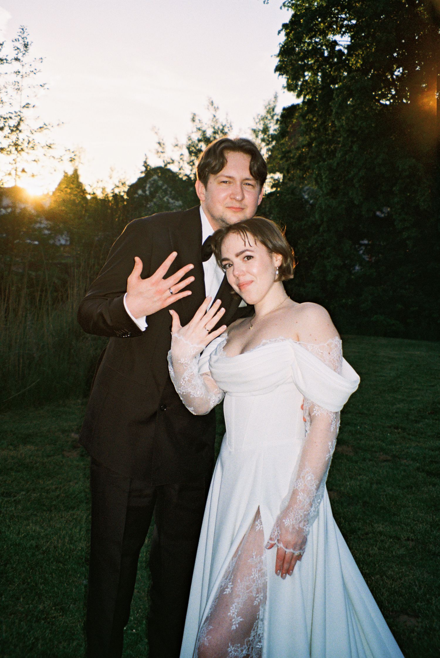 Couple in wedding attire posing at sunset outdoors, bride in off-shoulder lace gown, groom in black suit.