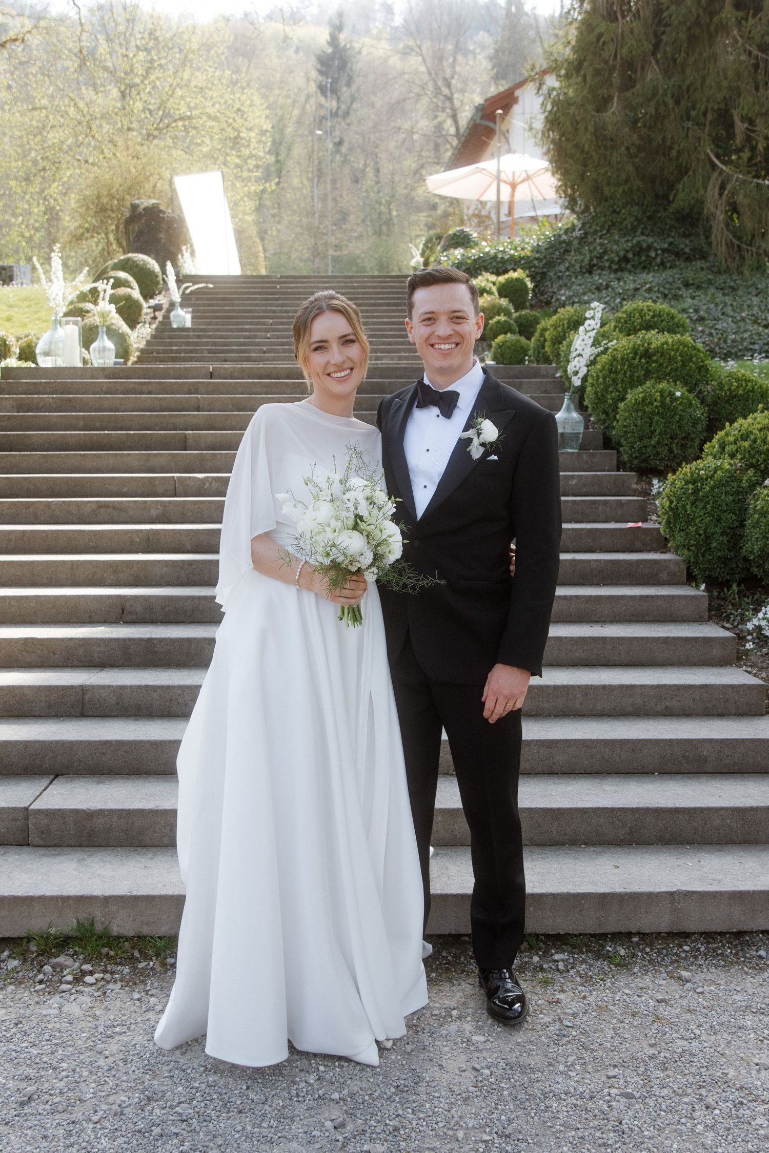 Bride and groom posing on stone steps in a garden, bride holding white bouquet, groom in black tuxedo with bow tie.