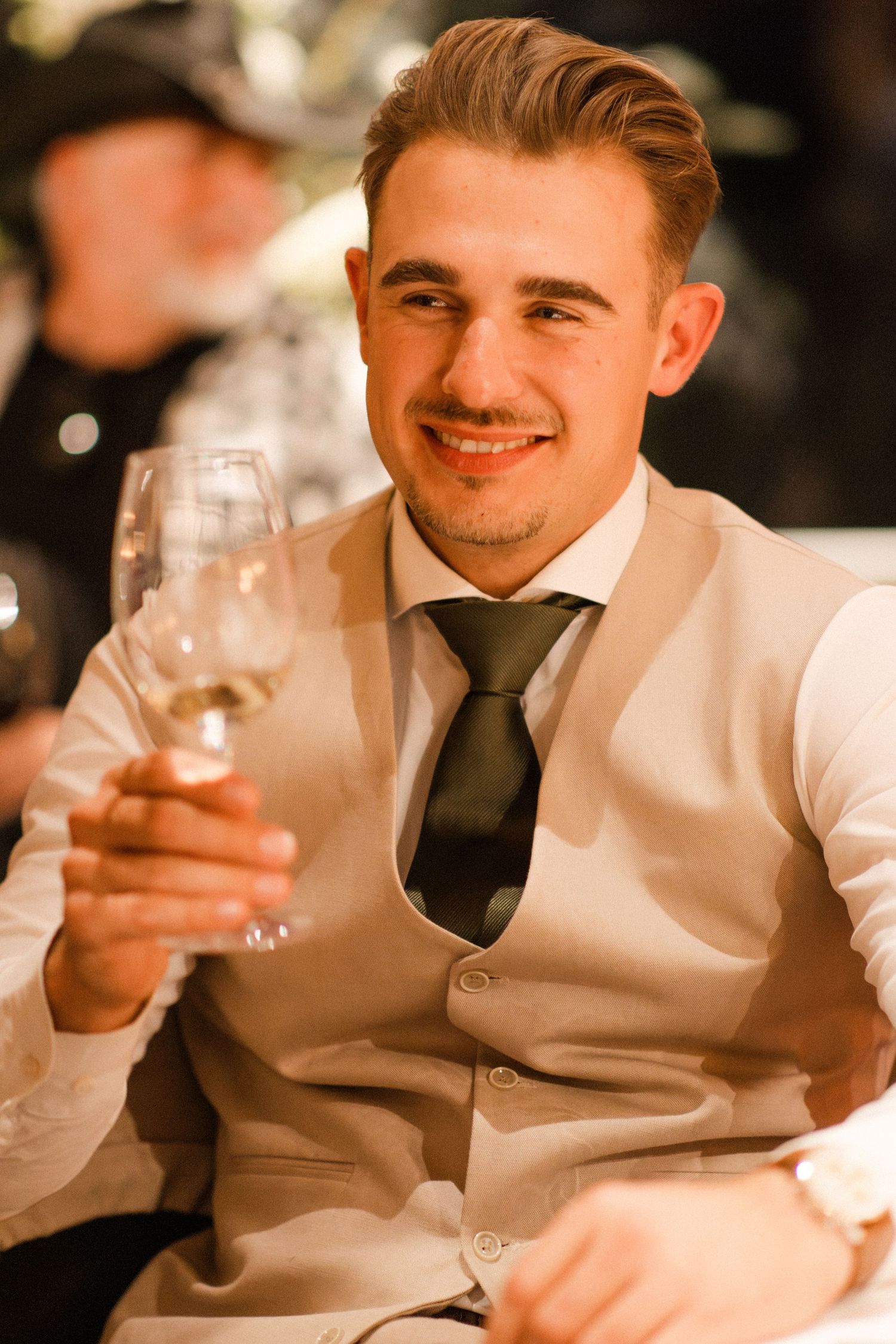 Smiling young man in vest and tie raising a wine glass at a formal event.