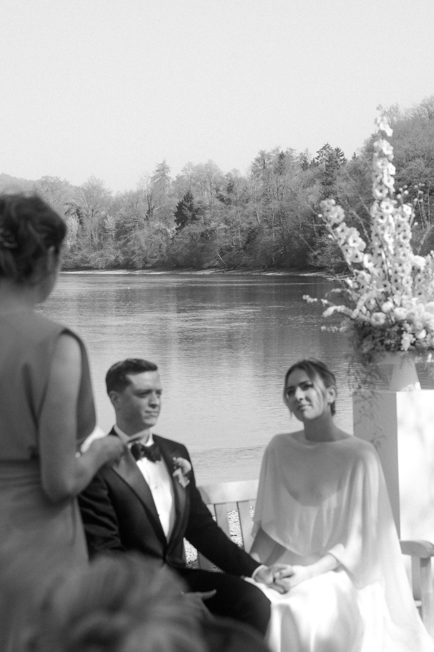 Black and white photo of bride and groom holding hands during lakeside wedding ceremony.