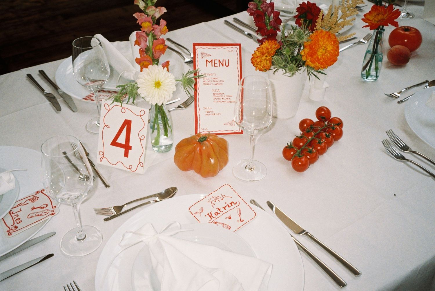 Elegant wedding table setting with white linens, floral centerpieces, cherry tomatoes, menu card, and number 4 table marker.