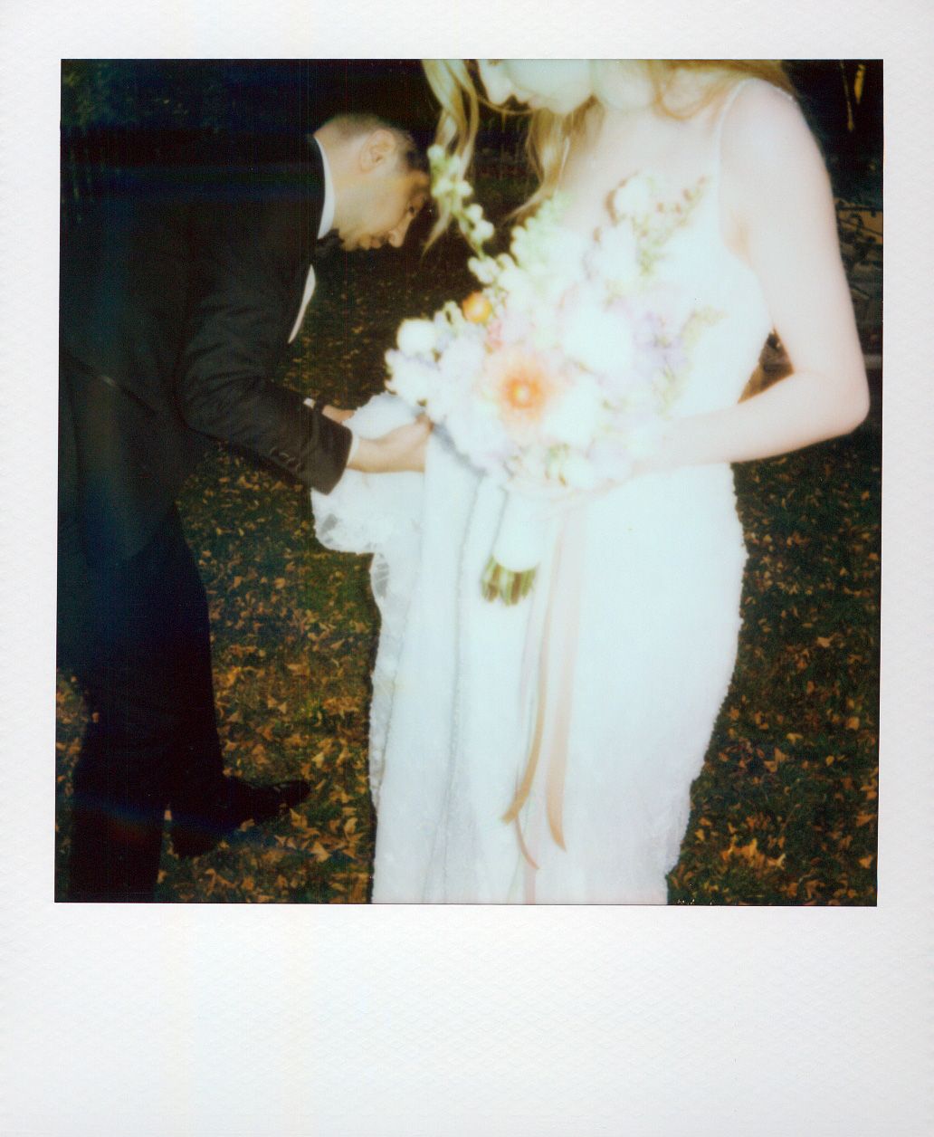 A person in a flowing white dress poses in front of scattered autumn leaves on the ground at dusk.