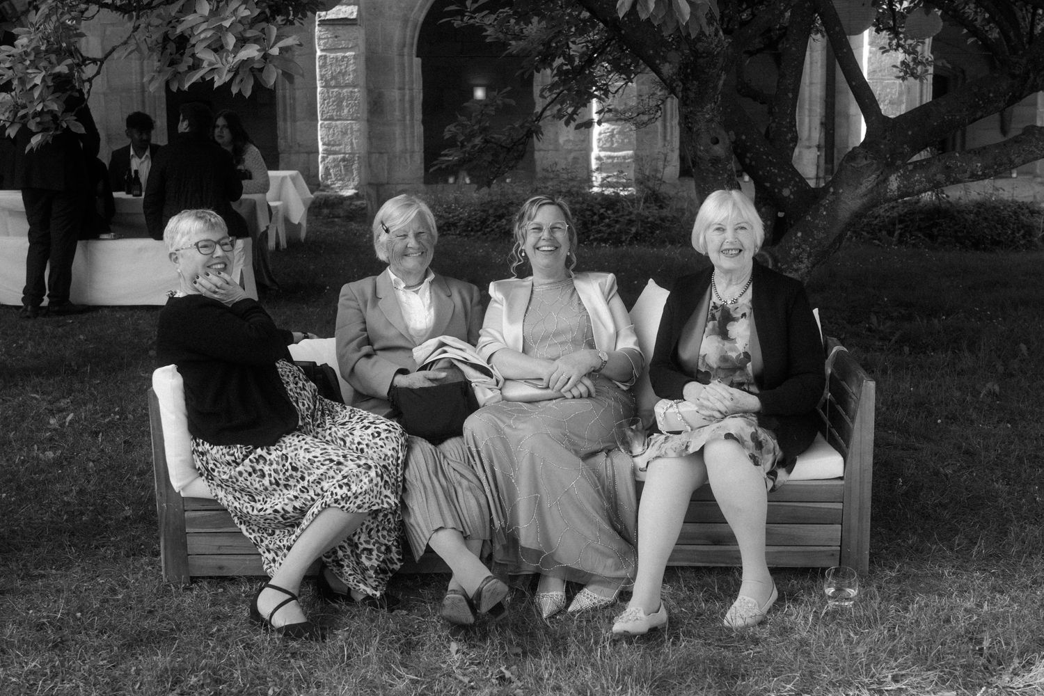 Wedding guests sit together on a bench in a garden setting.