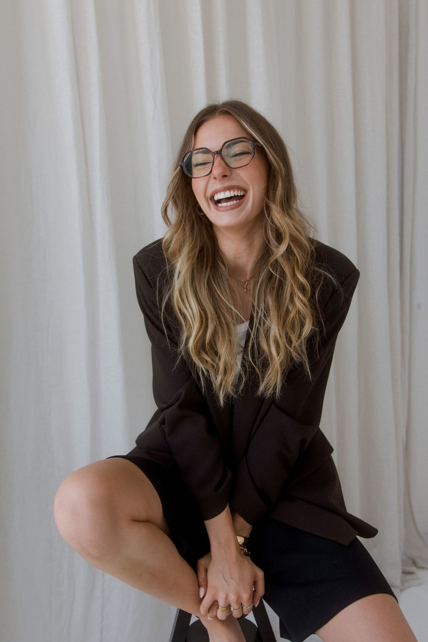 Smiling woman with wavy blonde hair and glasses wearing a dark blazer, seated casually against a white curtain backdrop.