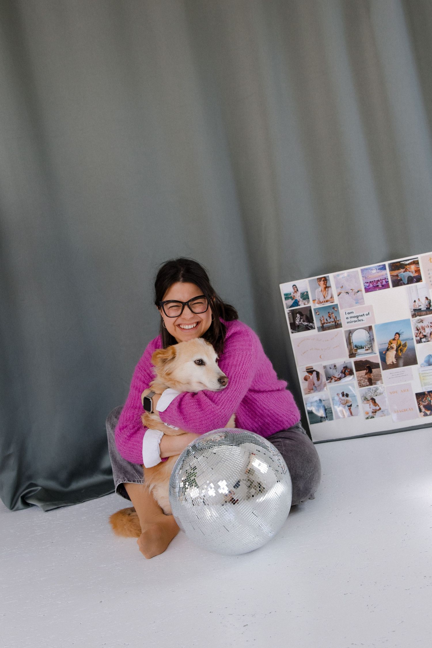 Woman in pink sweater hugging small dog beside disco ball and photo collage board on white floor.