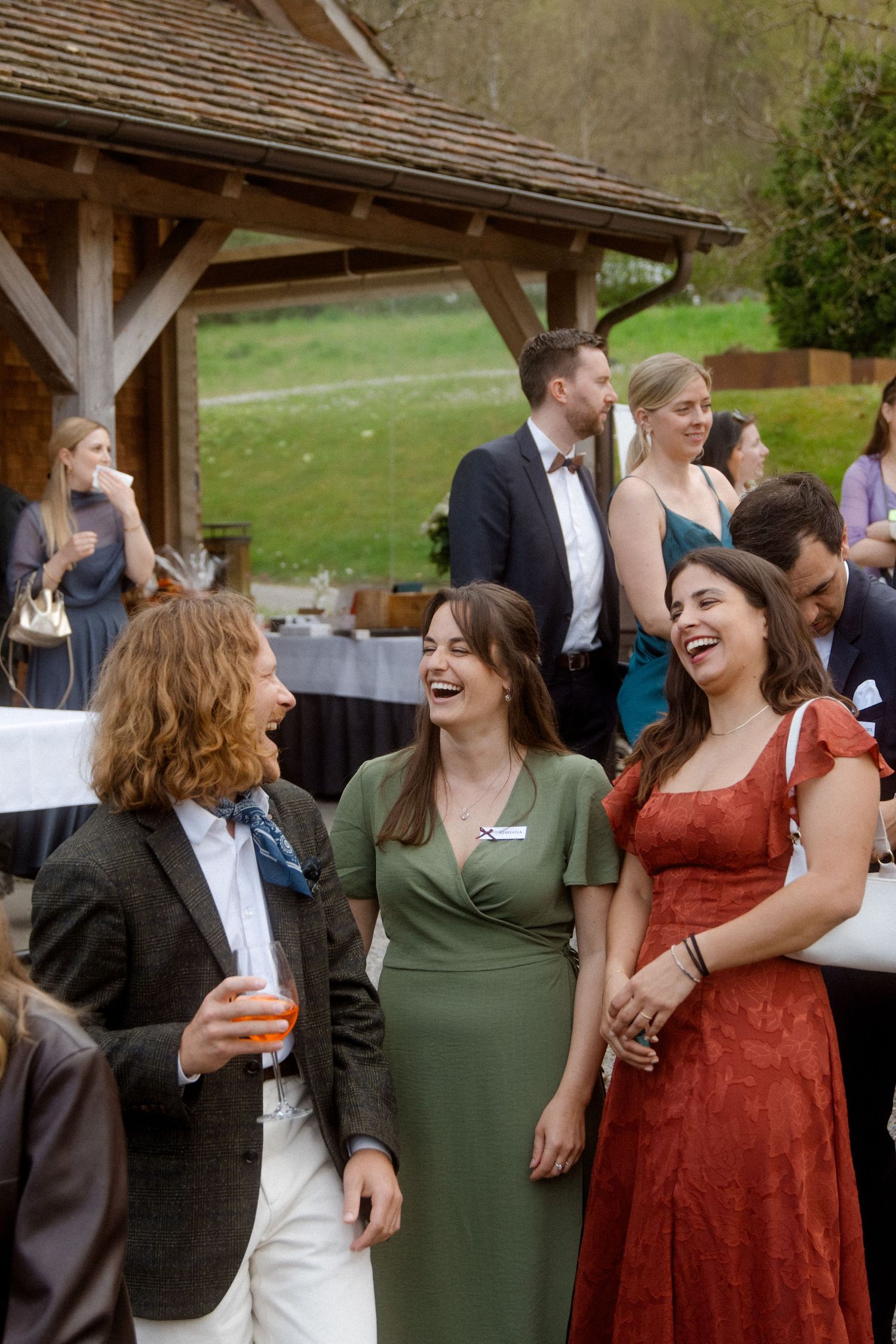 Wedding guests laughing together outdoors near rustic wooden pavilion during reception.