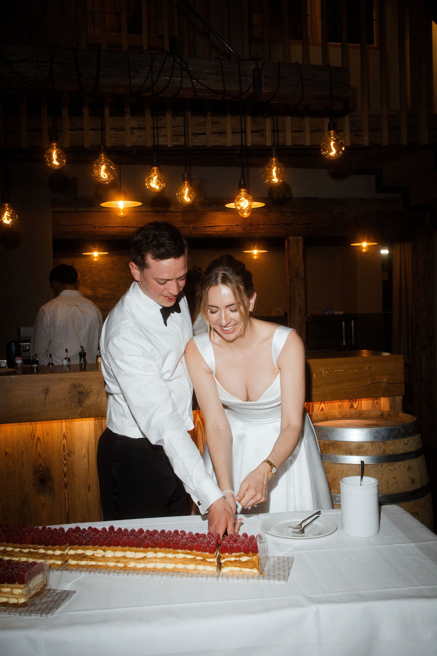 Bride and groom cutting wedding cake together in warmly lit rustic barn venue with Edison bulb lighting.