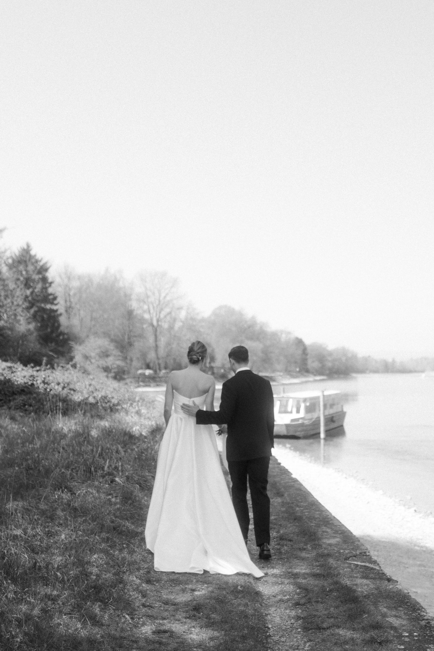 Black and white photo of a bride and groom walking hand in hand along a serene lakeside path on their wedding day.
