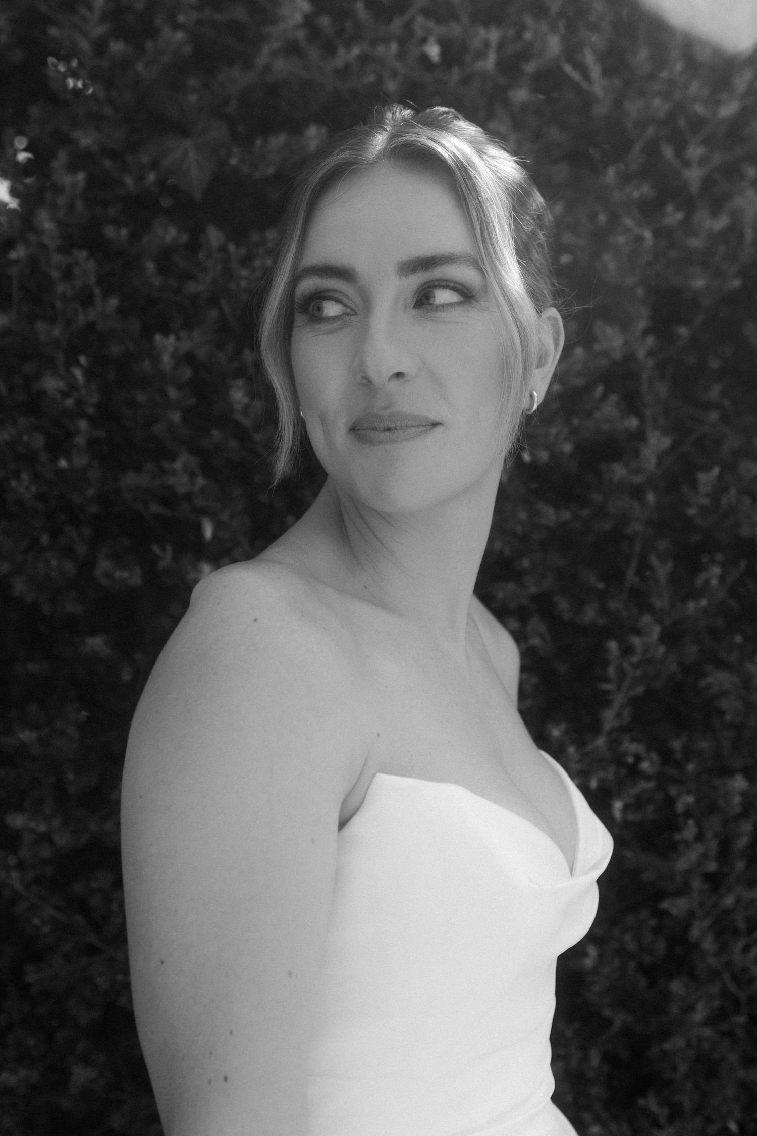 Black and white portrait of bride in strapless gown gazing away, green hedge background.