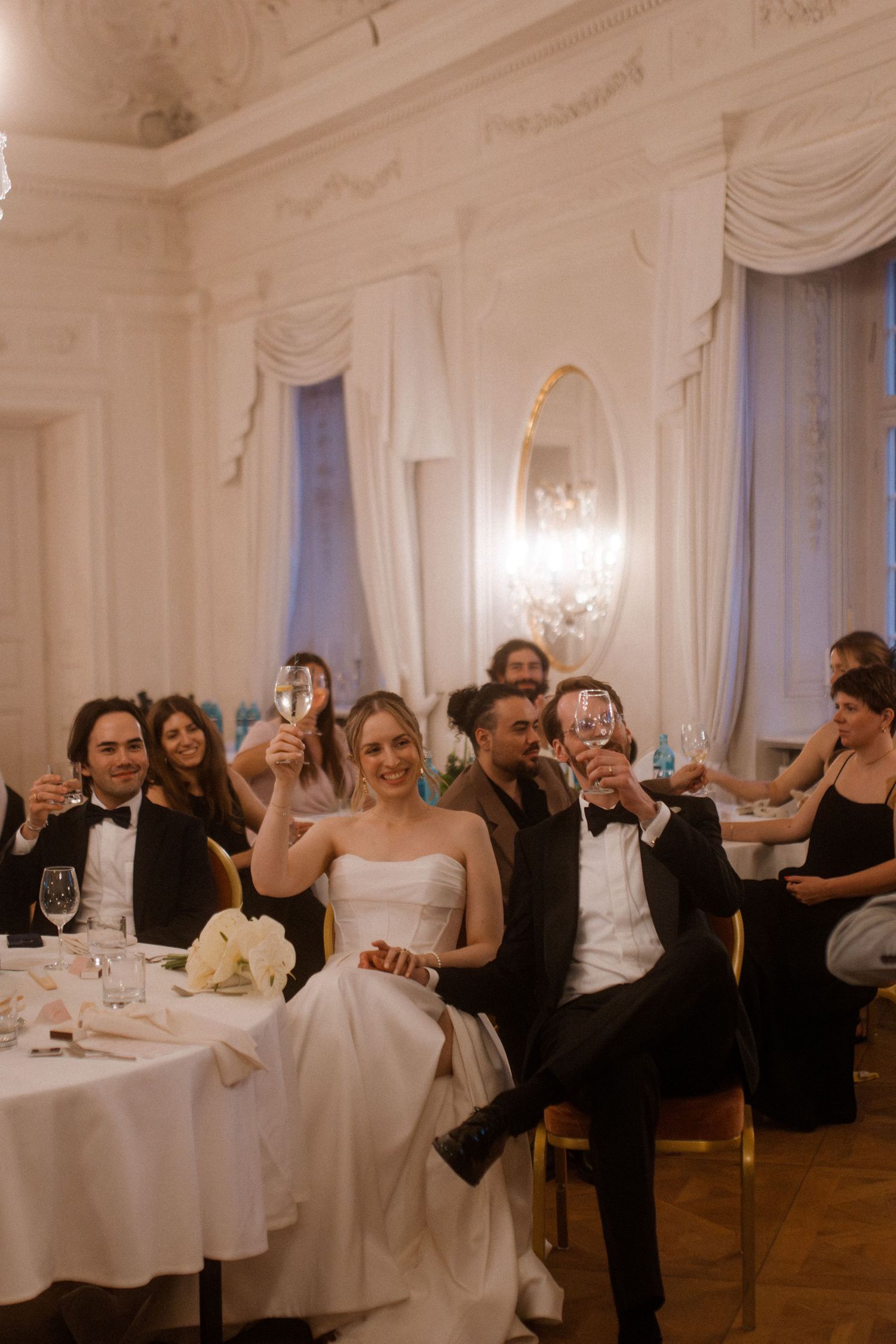 Wedding guests laugh and celebrate at round tables in an ornate white ballroom with chandeliers.