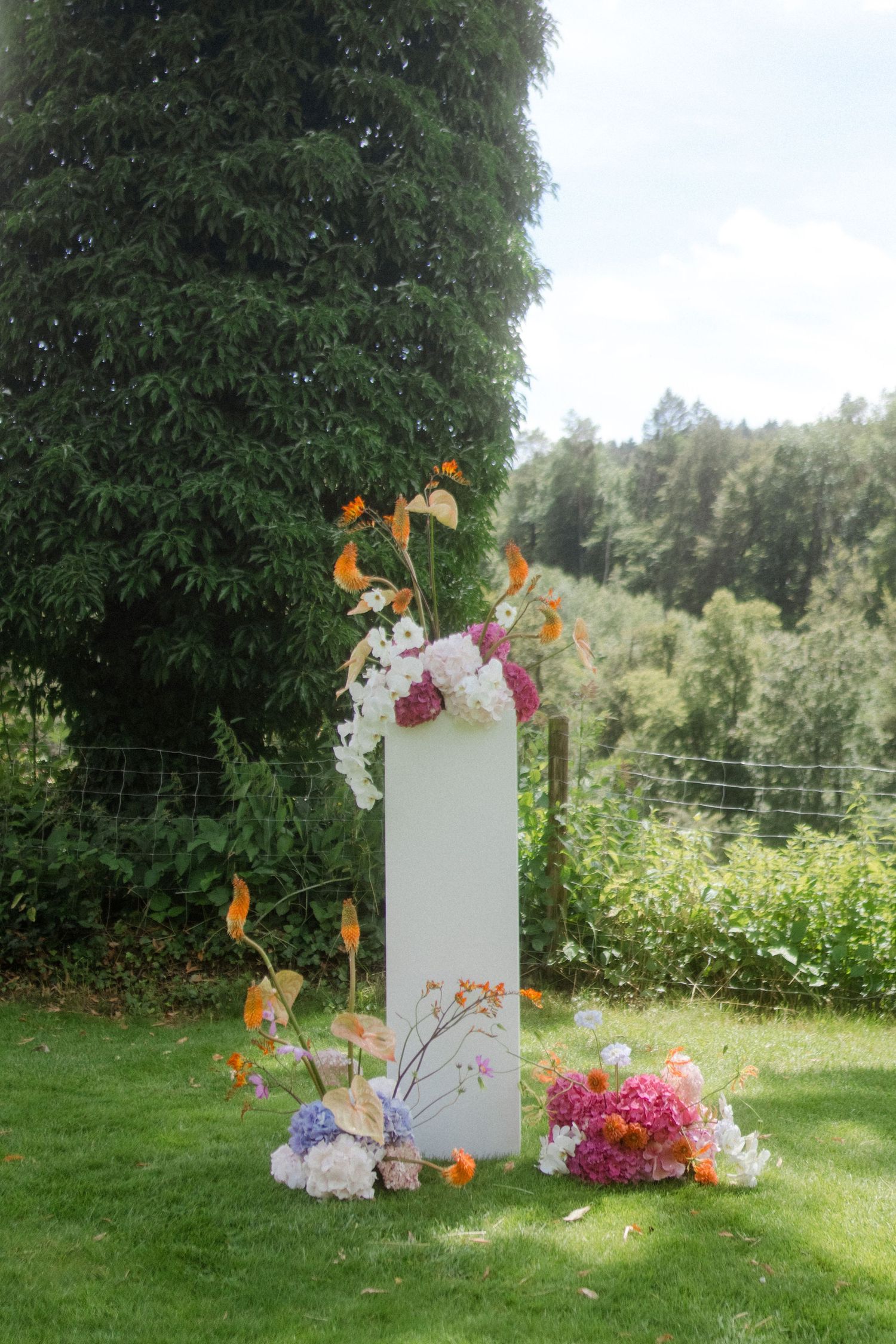White decorative pillar adorned with colorful flowers and greenery in outdoor garden setting.
