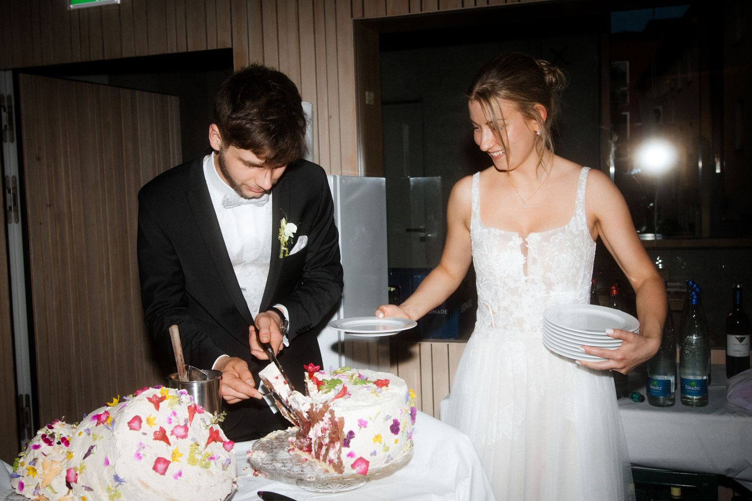A couple serves slices of their flower-adorned wedding cake at the reception.