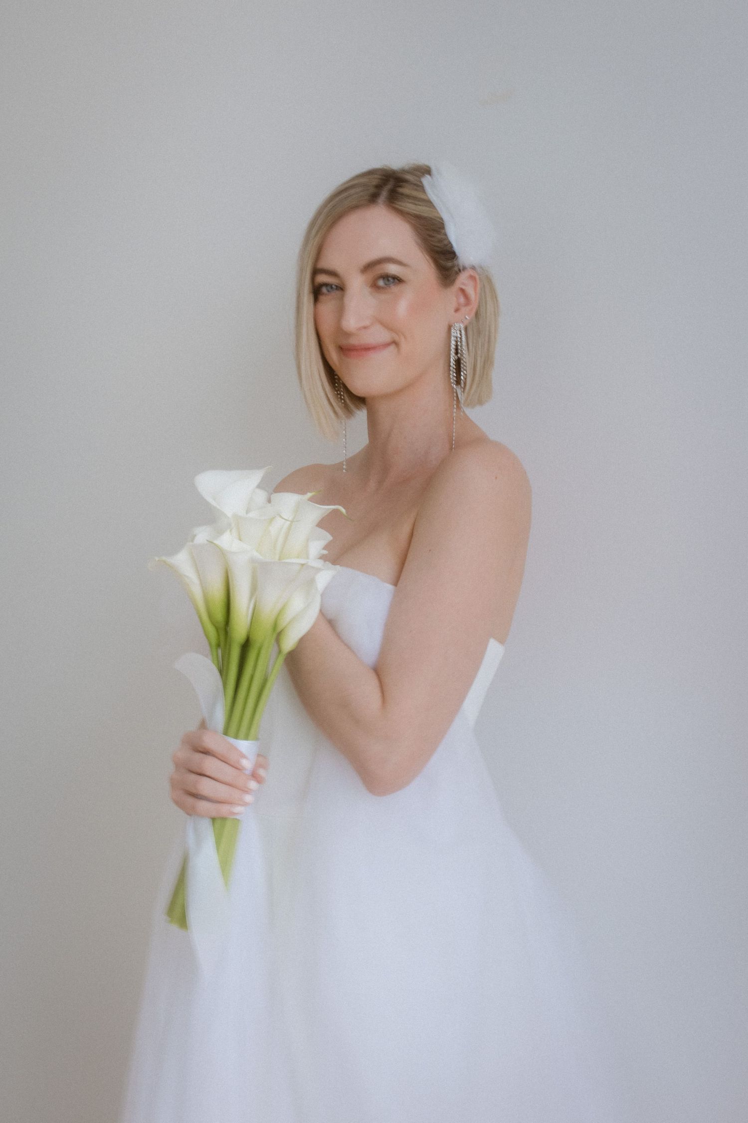 Bride in white wedding dress holding elegant calla lily bouquet against grey background.