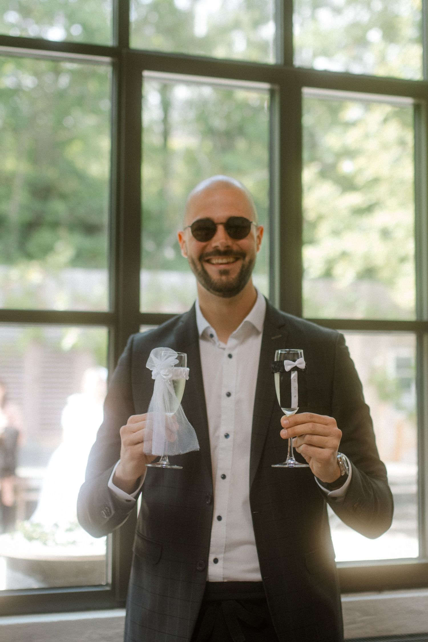 Wedding guest in a dark suit holds up two champagne glasses while smiling.