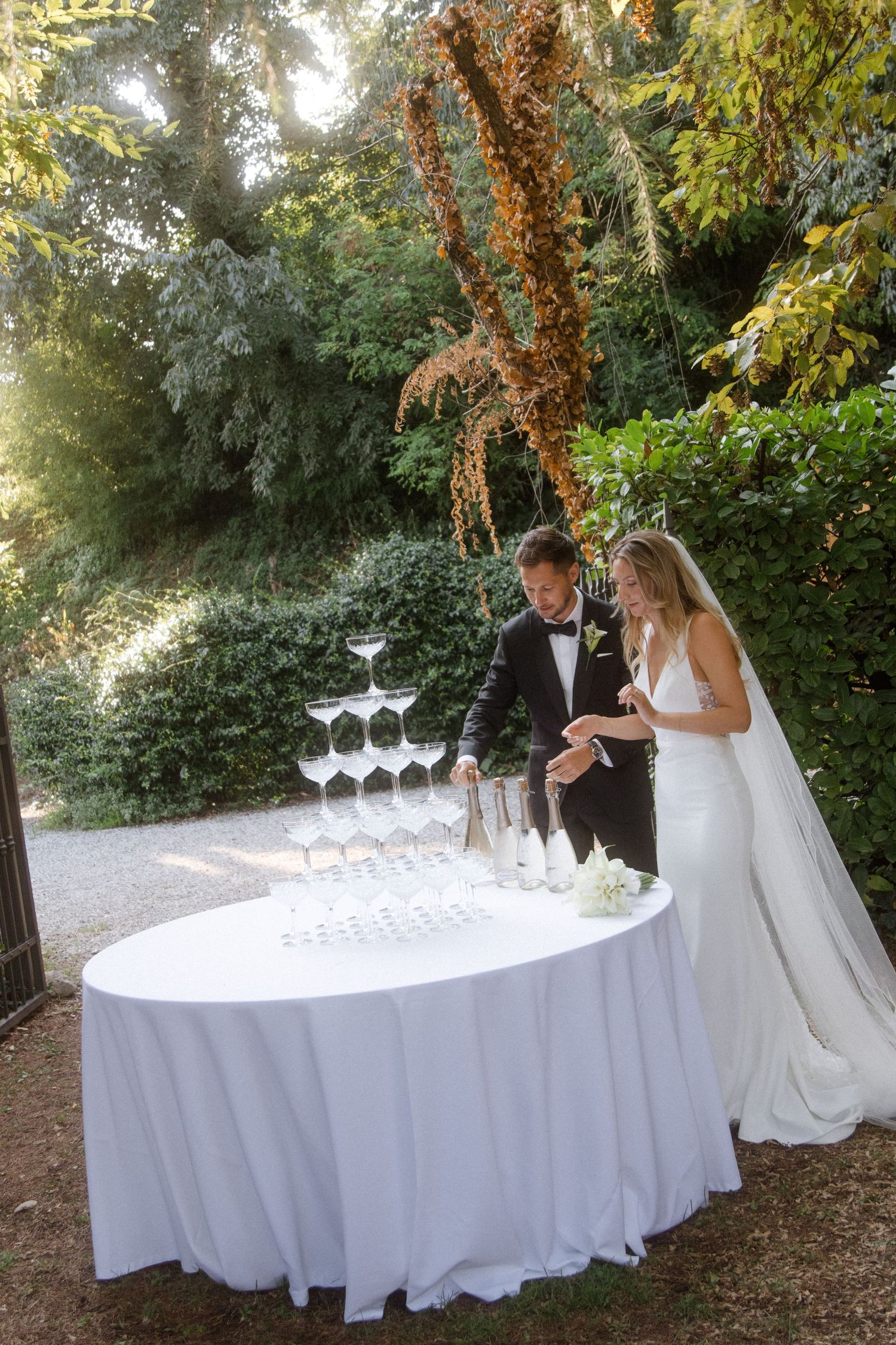 Newlyweds pouring champagne into a tower of glasses at an elegant outdoor garden reception.