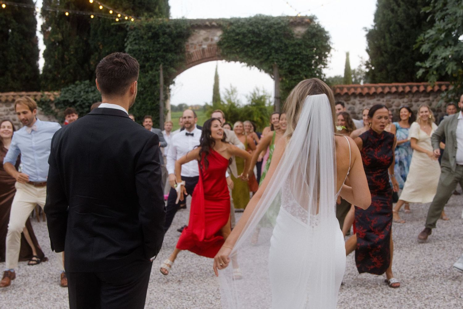 Wedding celebration with guests dancing under string lights and tall cypress trees.