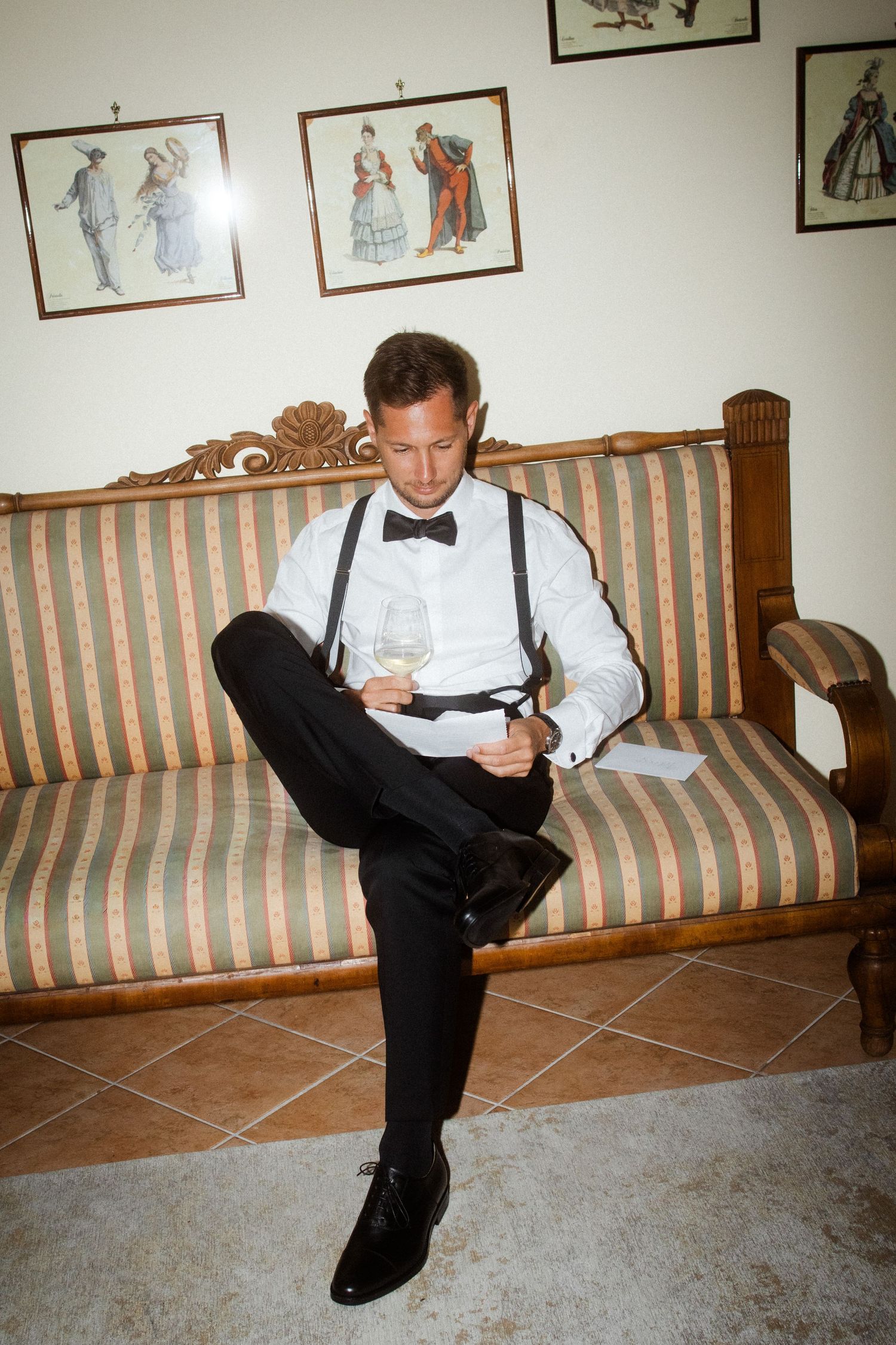 A person in formal wear reads a note while sitting on a striped vintage couch.