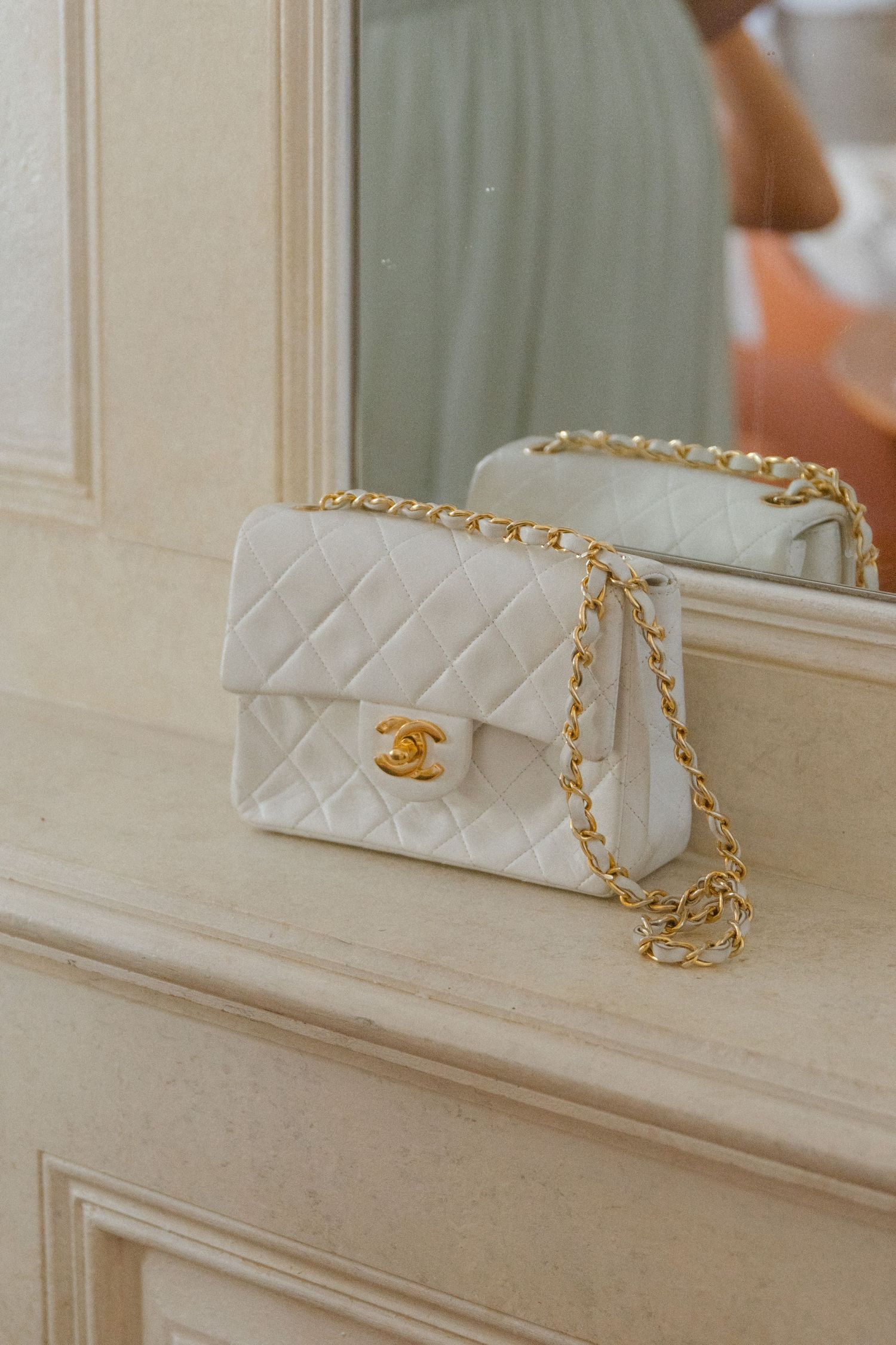White quilted Chanel handbag with gold chain strap displayed on a cream colored ledge.