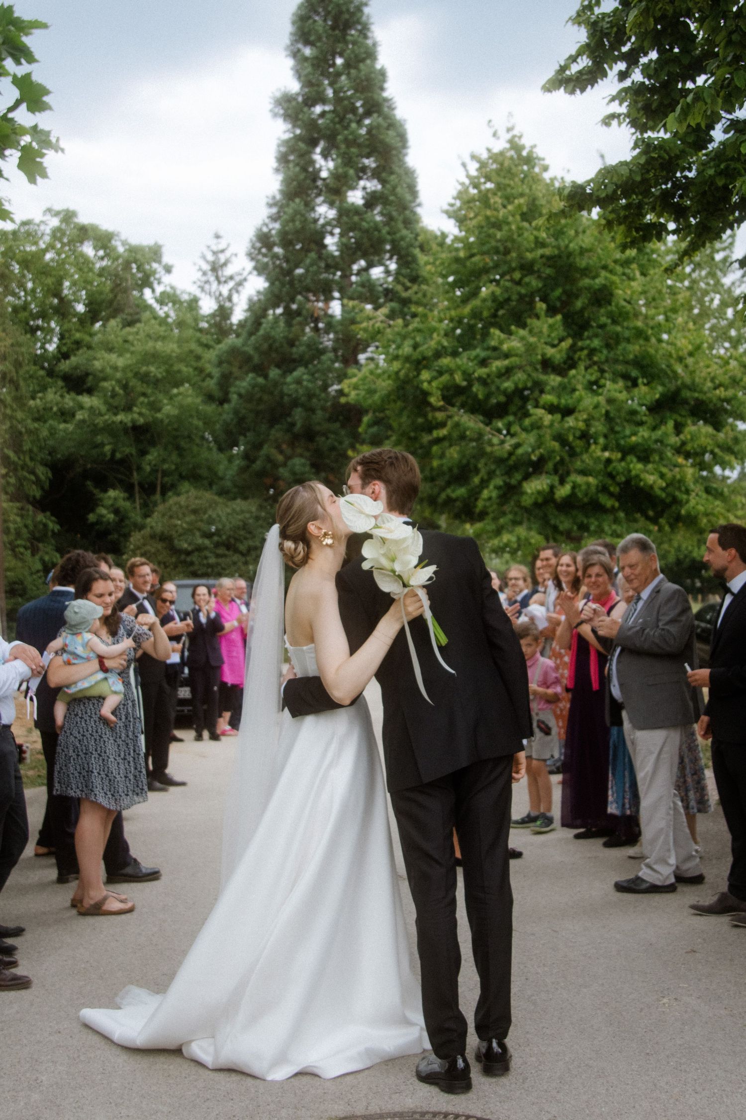 Wedding guests form a receiving line outdoors under green trees on a paved pathway.