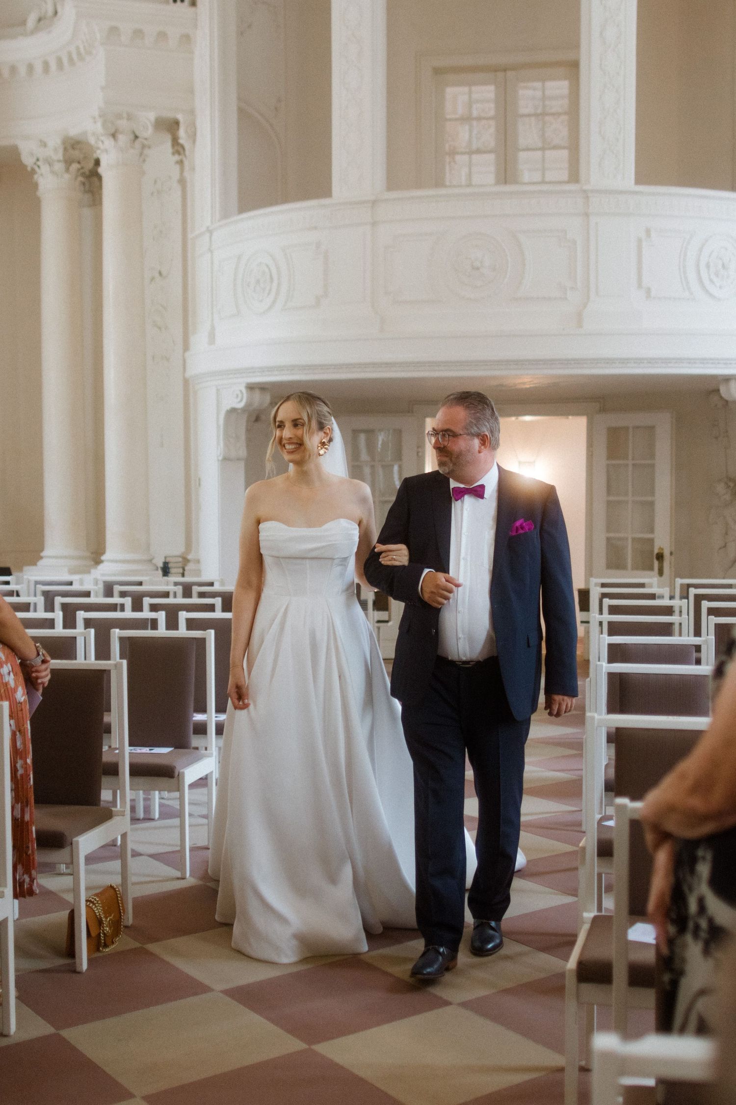 A couple walking down the aisle of a chapel with white columns and checkered floor.