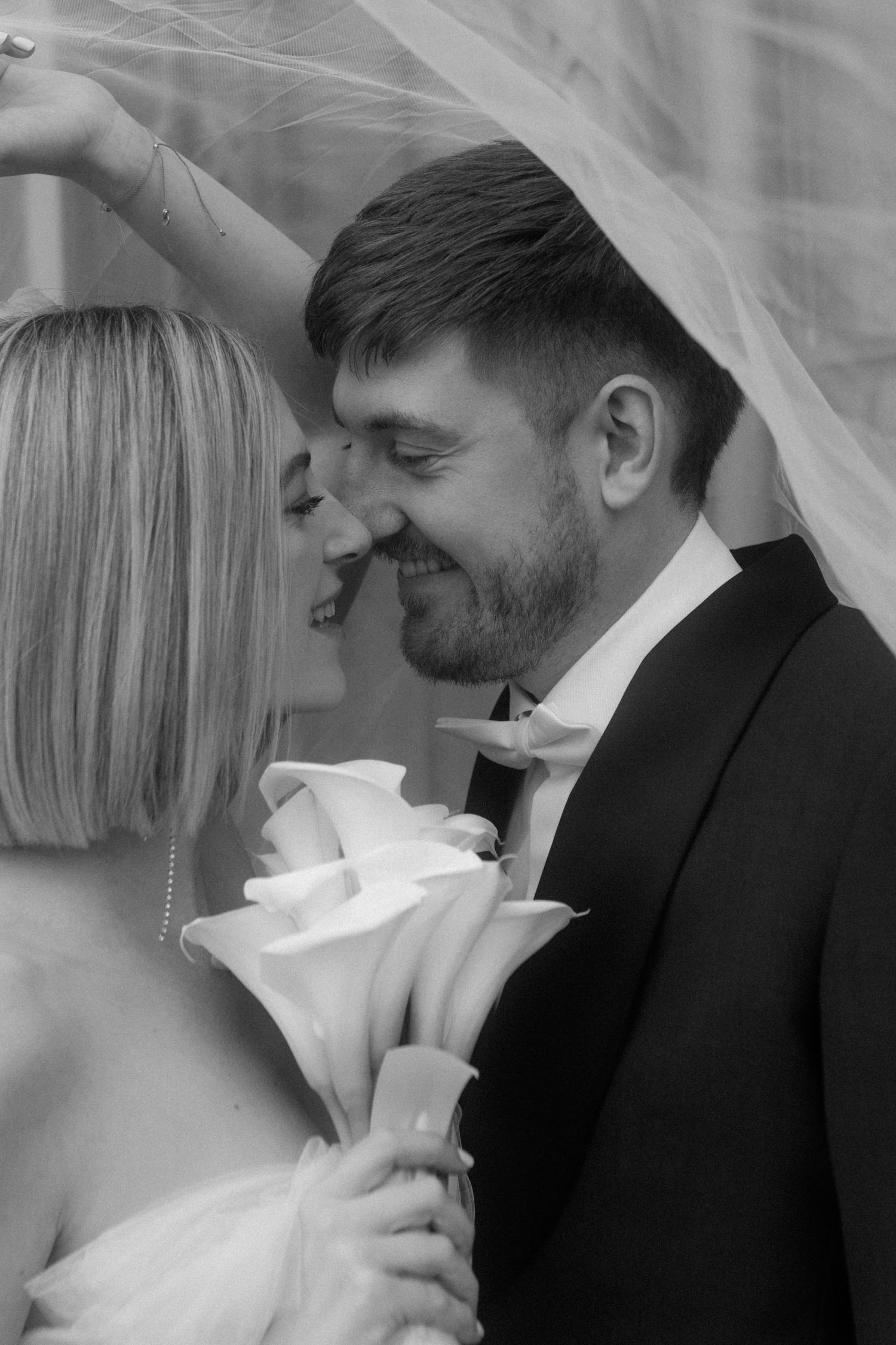 Romantic black and white photo of newlyweds sharing an intimate moment under a veil with white calla lilies.