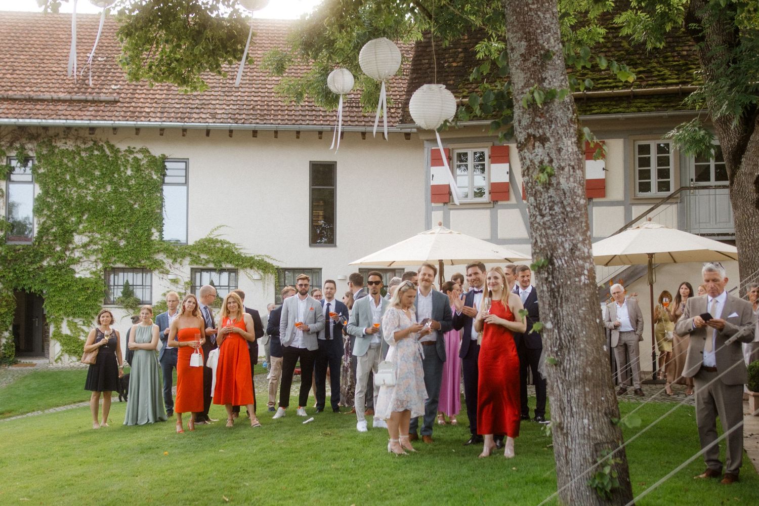Wedding guests gather on a grassy lawn decorated with white lanterns during an outdoor celebration.
