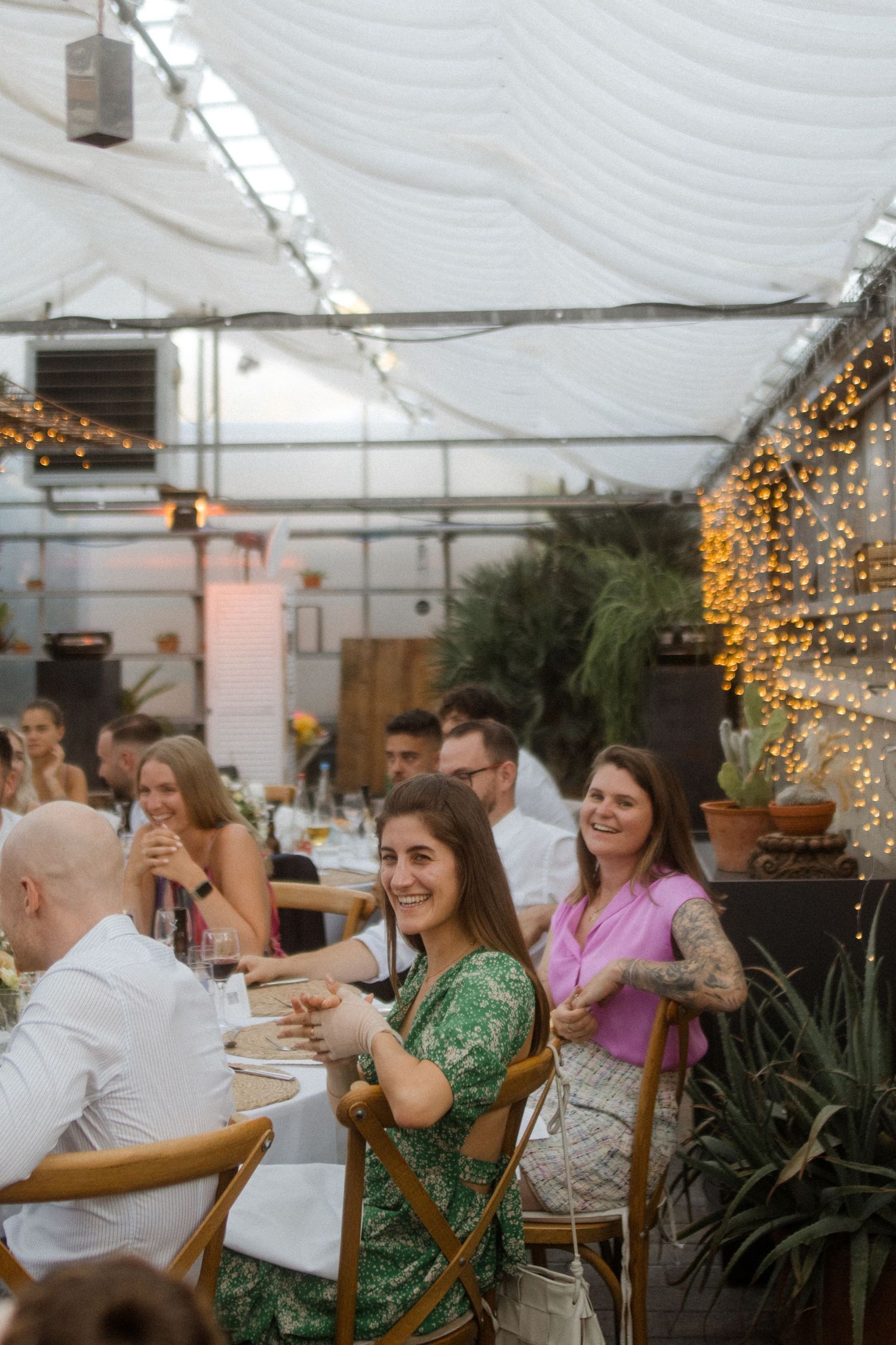 Wedding guests enjoying dinner under string lights in greenhouse venue.