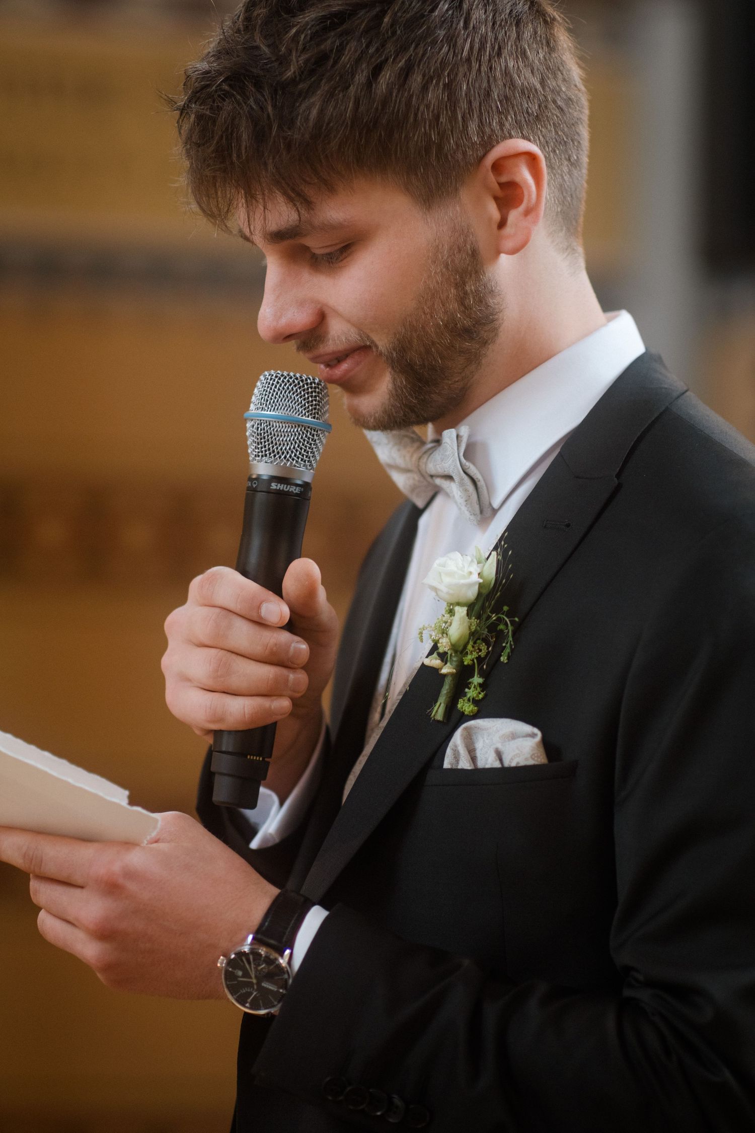 A groom reads from paper while speaking into microphone during wedding ceremony.