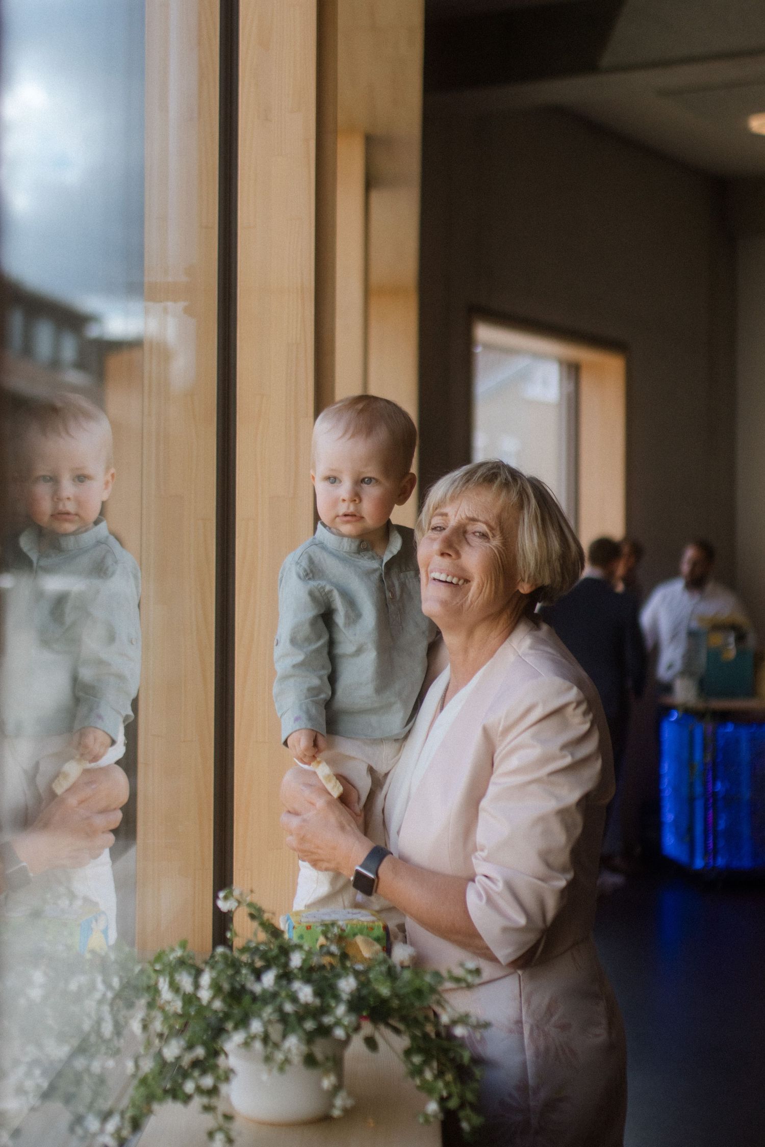Woman in beige jacket joyfully interacts with small child by window with potted flowers.