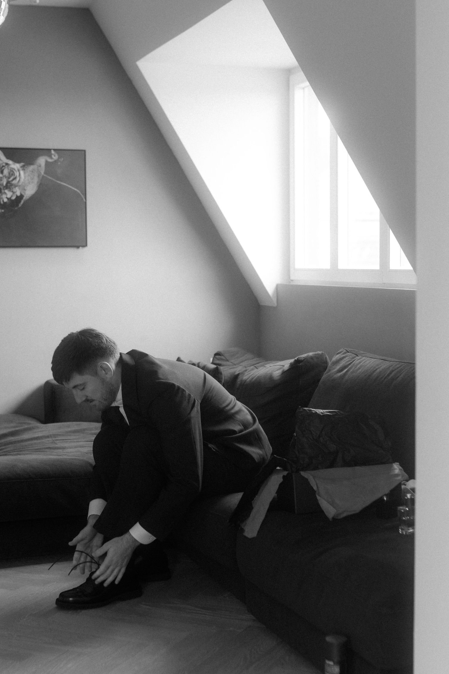 Person in dark suit adjusting shoes while seated on a couch under slanted window.